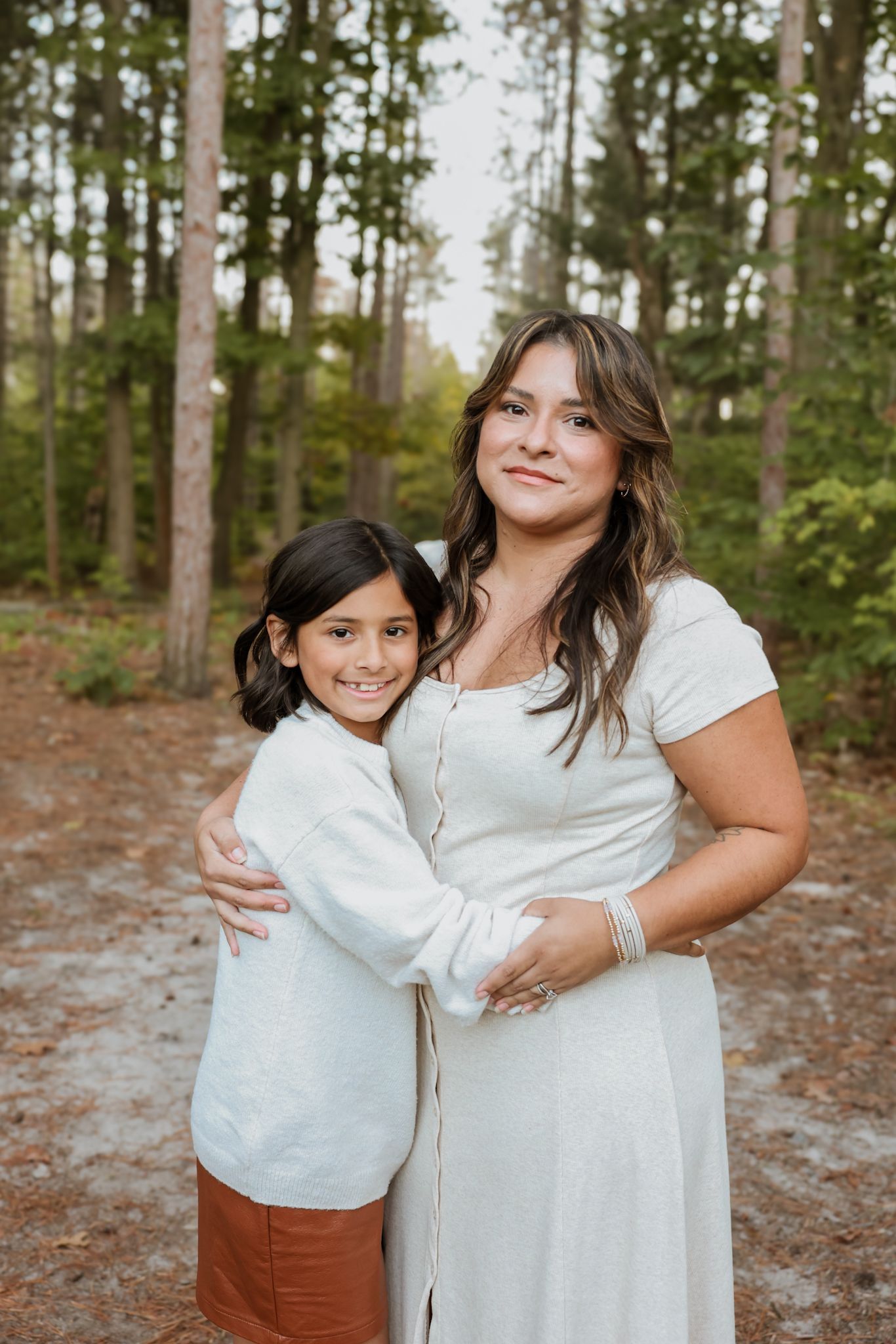 Older daughter in white sweater hugs pregnant mother in white dress in pine forest.