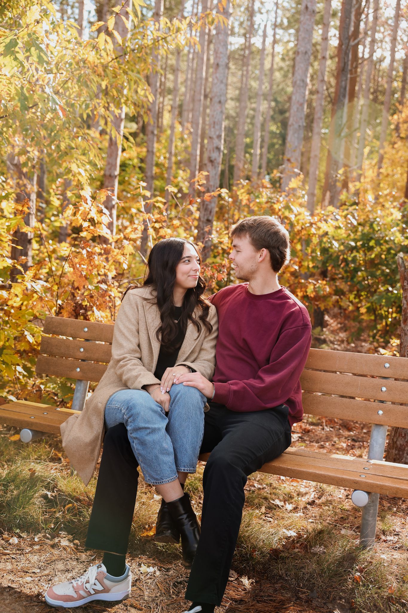 A smiling couple sits closely on a wooden bench in a sunlit autumn forest, gazing at each other.