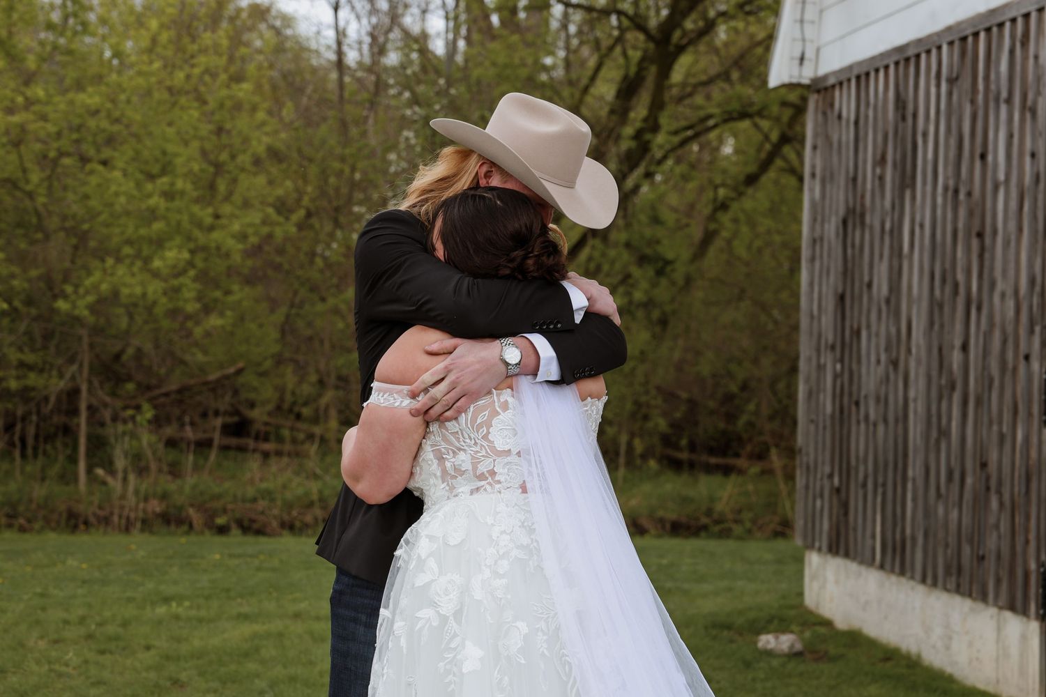 Bride in lace wedding gown and groom in cowboy hat share an emotional first look hug outdoors.