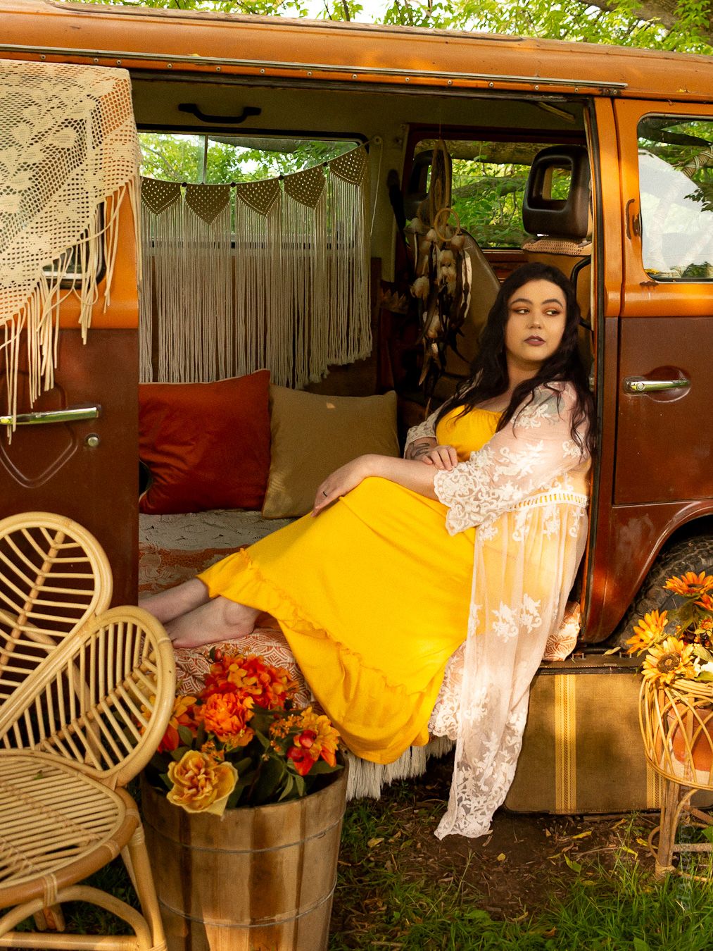 Woman in yellow dress and lace robe lounging in decorated vintage brown van outdoors.