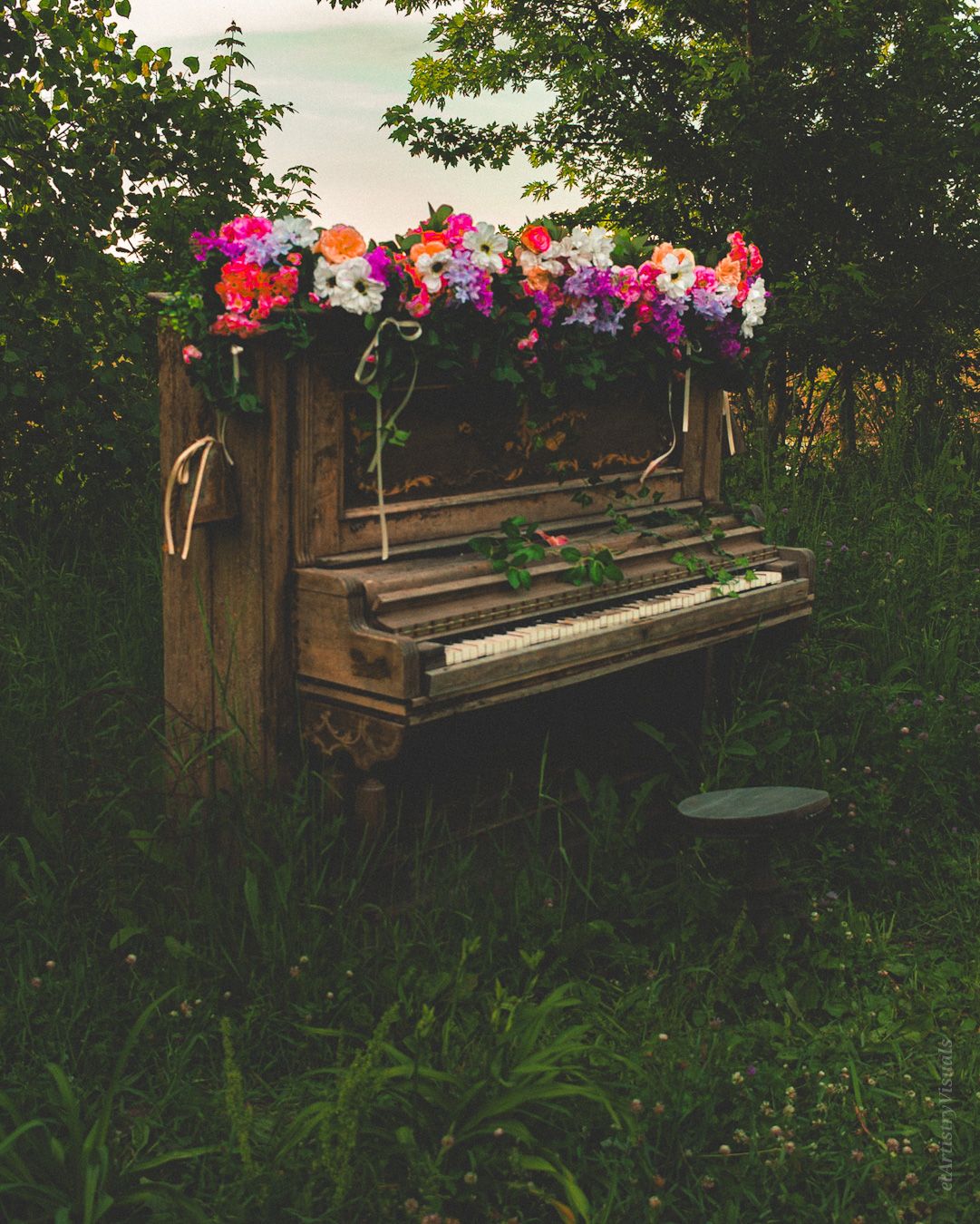 An old rustic upright piano decorated with colorful flowers sits outdoors surrounded by lush green foliage.