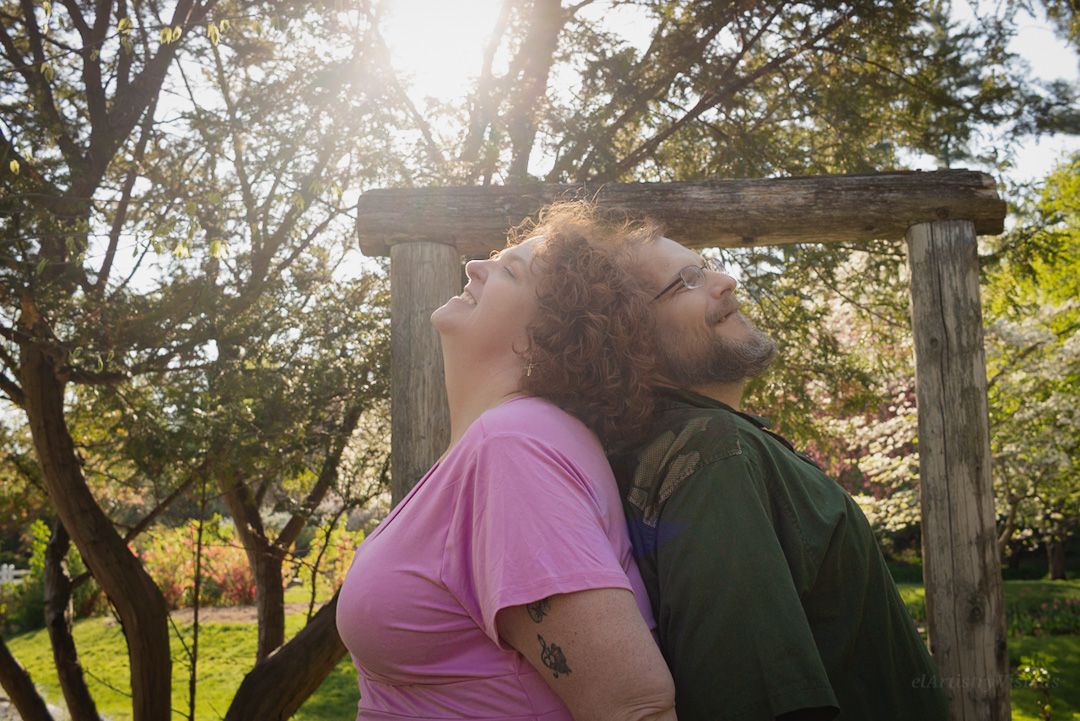 Couple leaning back-to-back under a wooden arbor surrounded by blooming trees in golden sunlight.