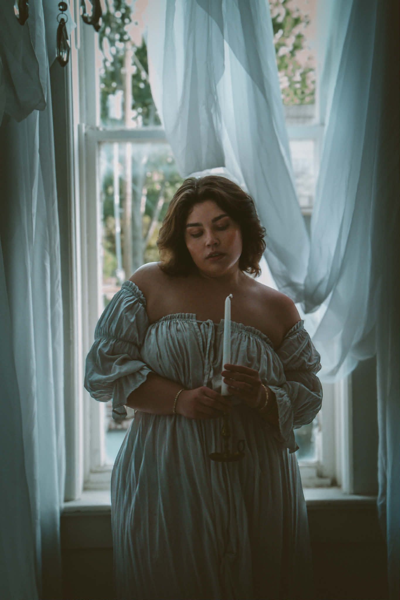 A woman in an off-shoulder blue vintage dress holds a candle near sheer white curtains by a window.