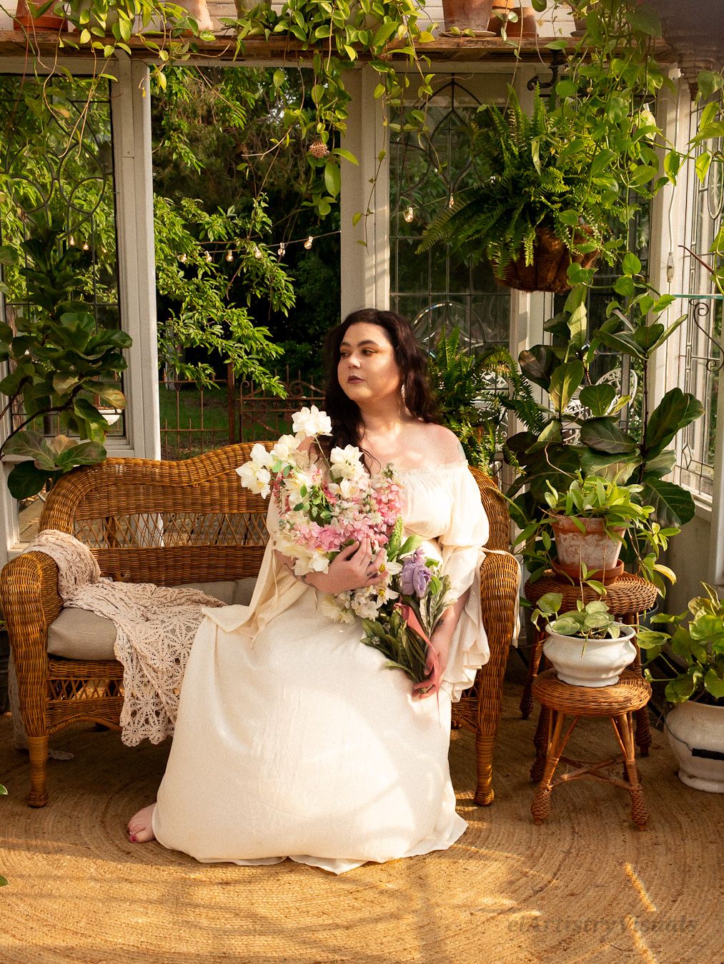 Woman in white gown holding bouquet seated on wicker sofa in lush indoor garden setting.