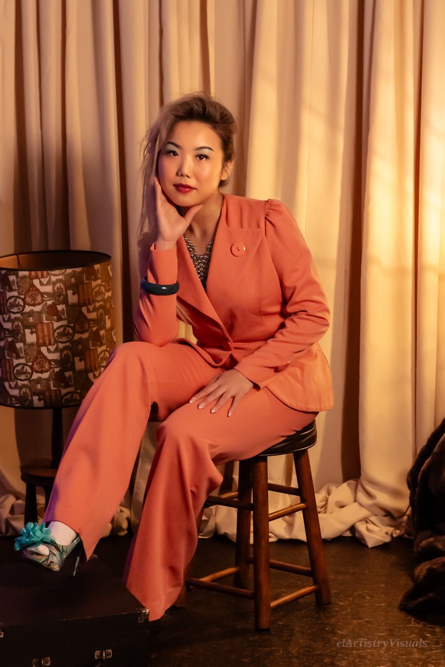 An Asian woman in an elegant coral pantsuit poses on a wooden stool in a warmly lit vintage room.