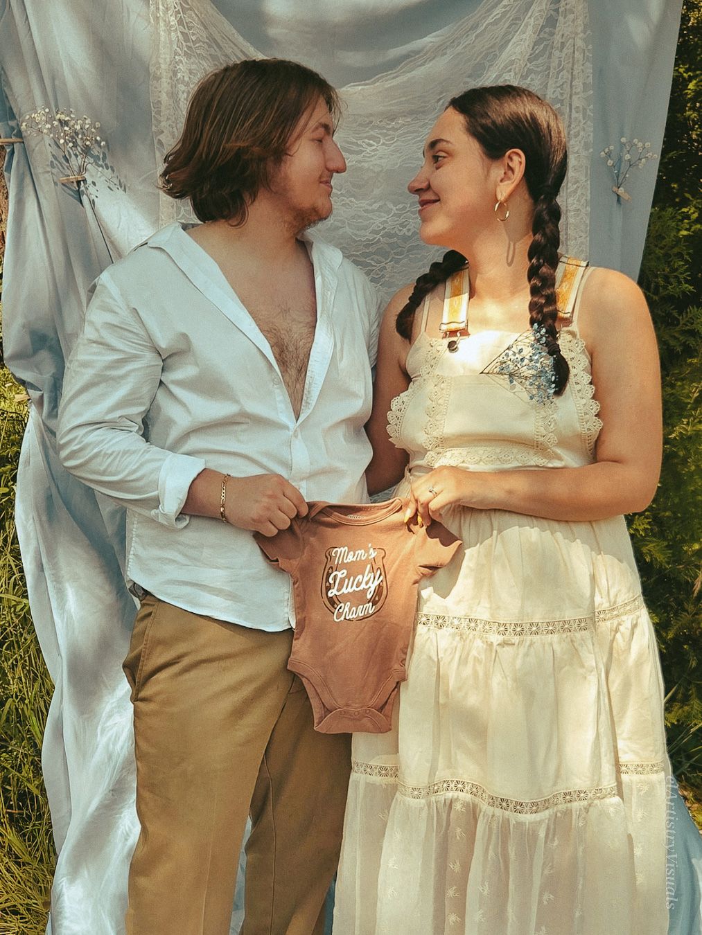A couple smiles at each other while holding a baby onesie during an outdoor maternity announcement photo shoot.