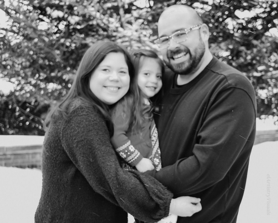 A smiling family of three shares a warm embrace in a black and white outdoor portrait photo.