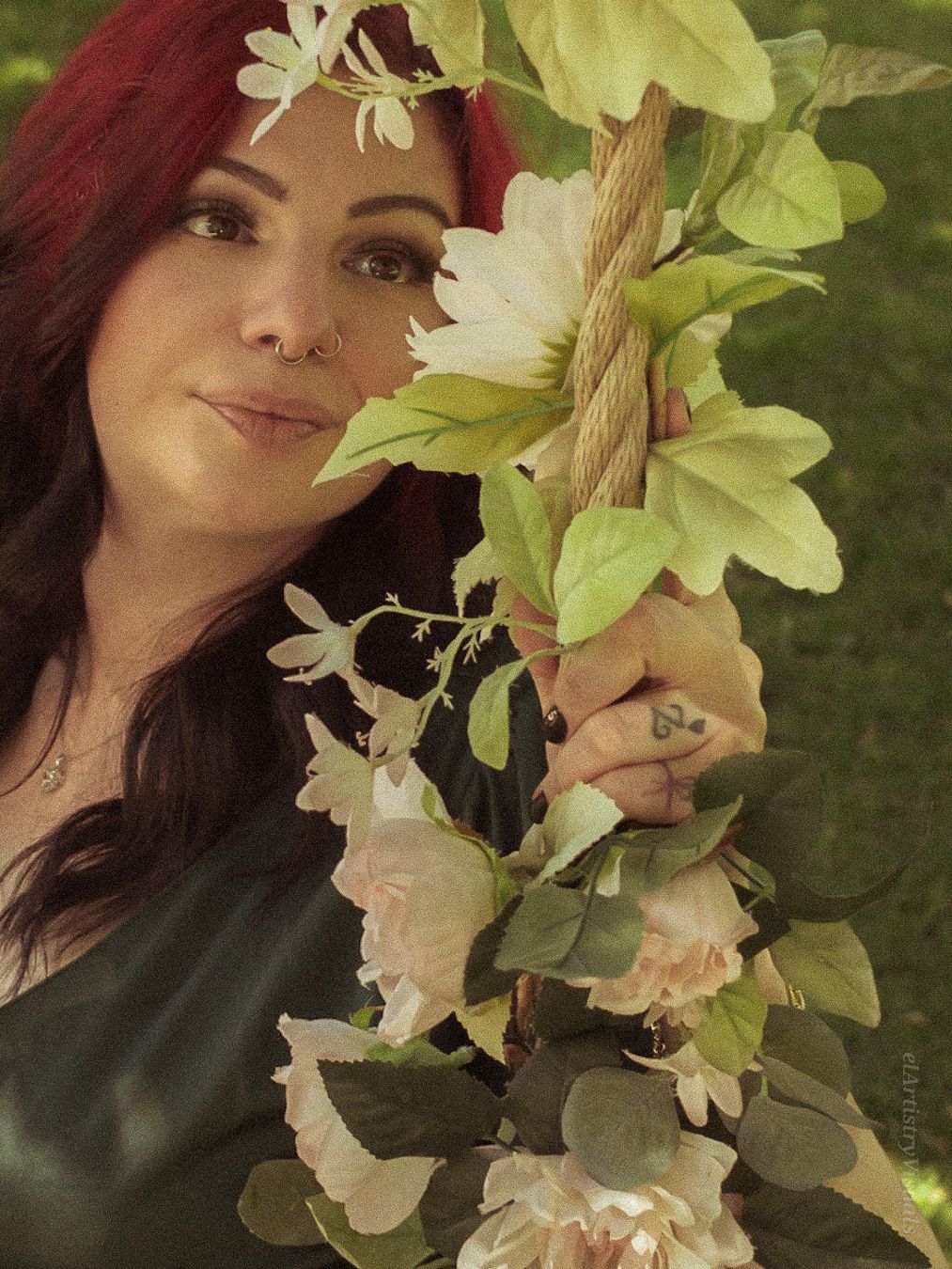 A woman with red-tipped dark hair smiles beside a floral rope swing with white blooms outdoors.