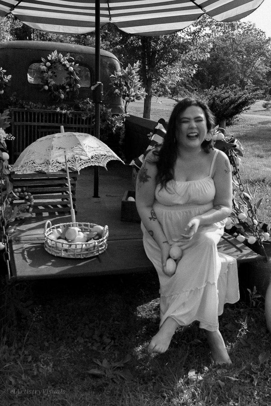 A laughing woman in a white dress sits outdoors at a vintage-styled photo shoot with an antique truck backdrop.