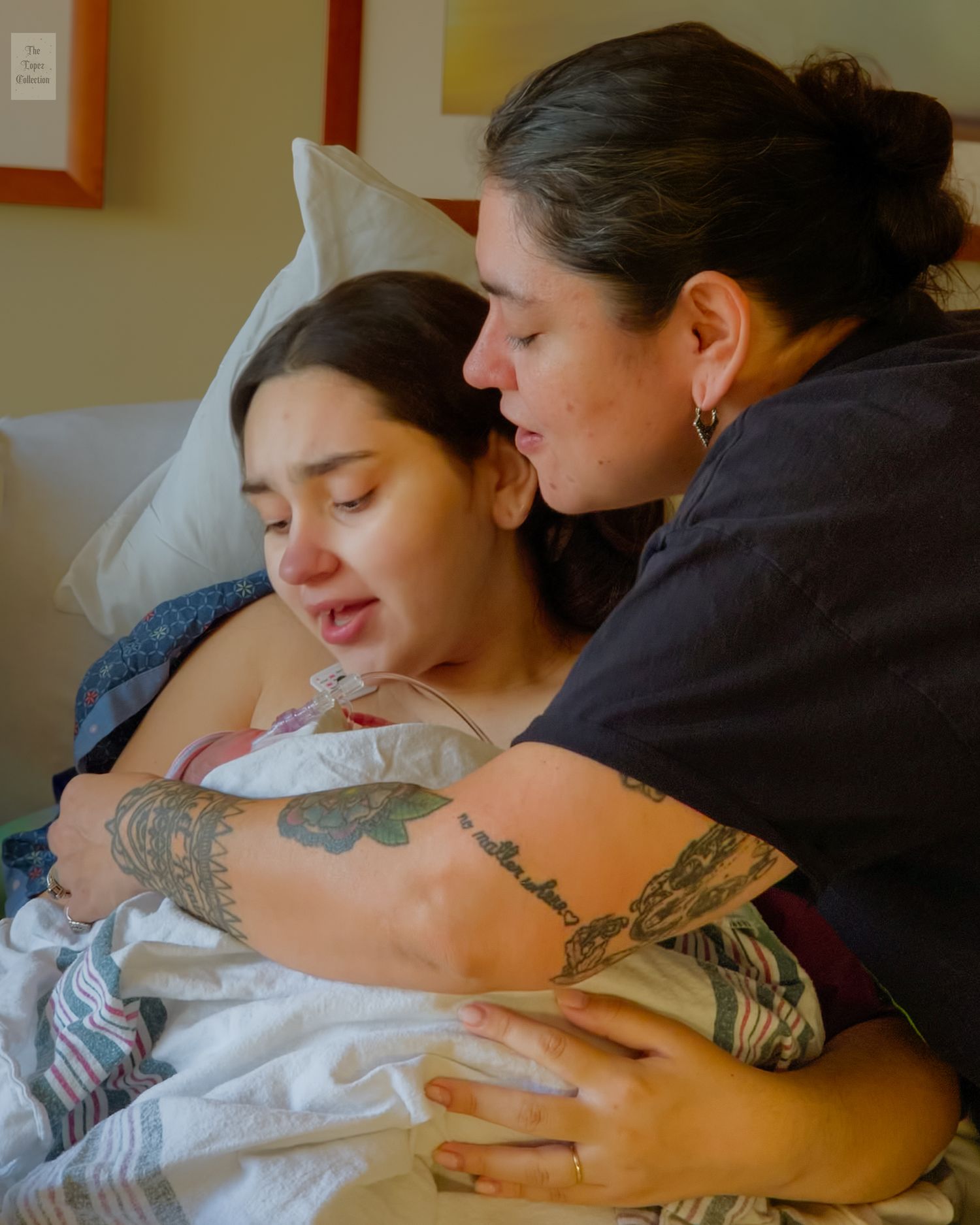 An emotional new mother holds her newborn while being comforted by a tattooed support person in a hospital room.