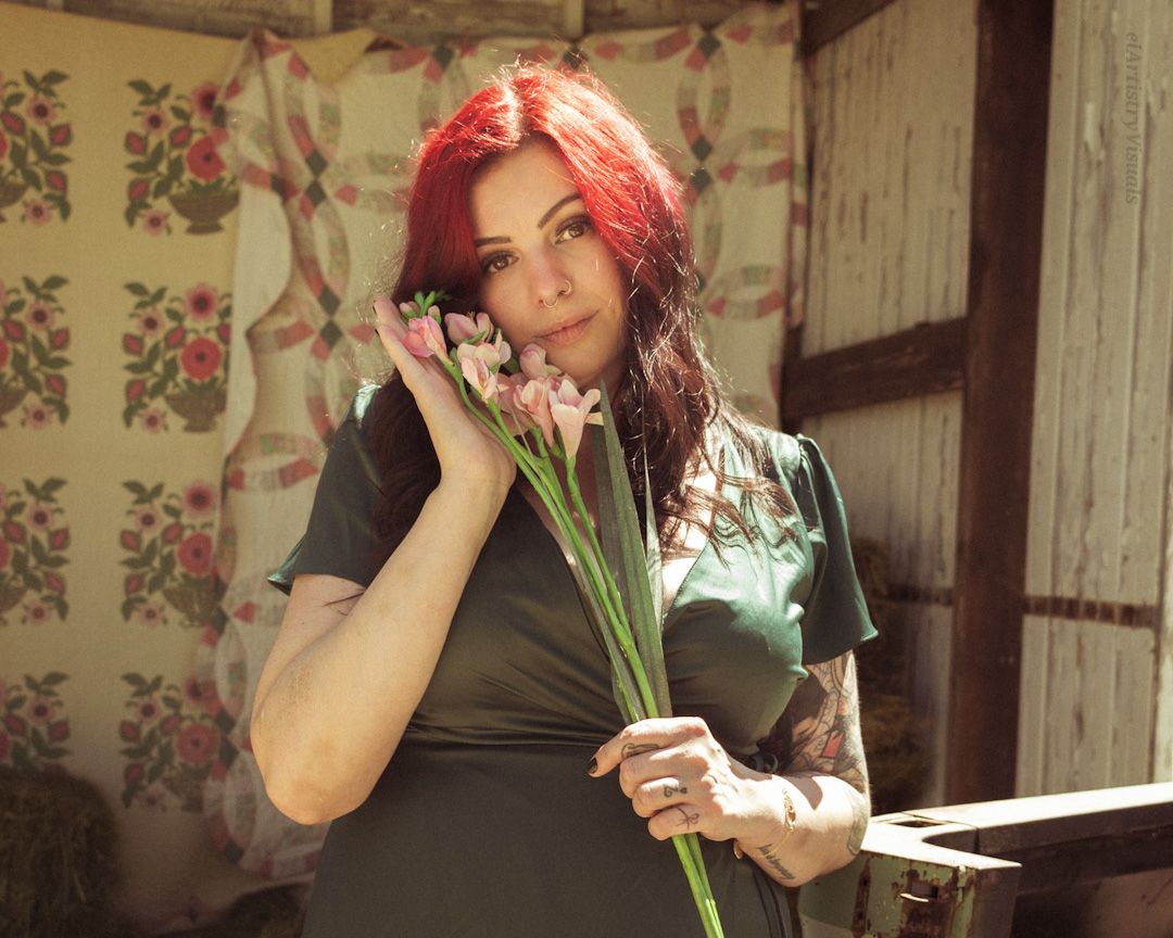 Red-haired tattooed woman in green dress holding pink flowers in rustic outdoor setting.