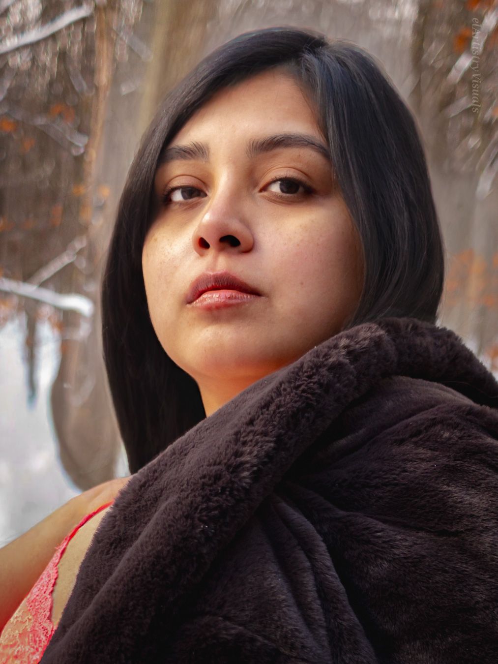 A young woman with dark hair wrapped in a dark fur coat poses outdoors in a winter forest setting.