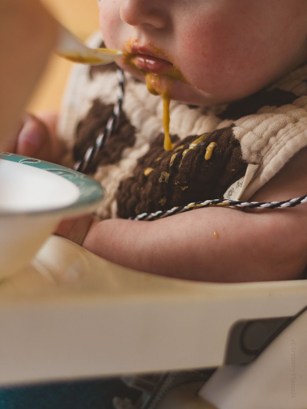 A baby in a high chair eating pureed food with a spoon, wearing a knit sweater bib.