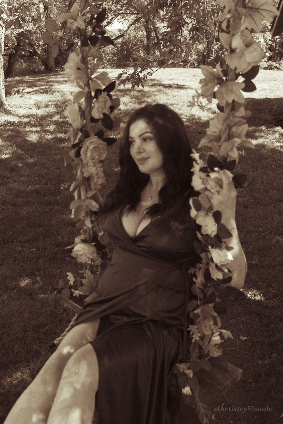 Woman in black satin dress sitting on floral swing in sepia-toned outdoor portrait.