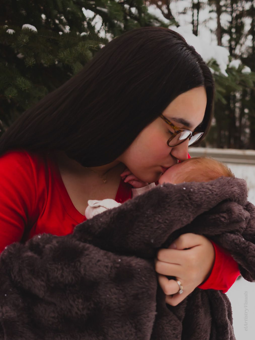 A mother in a red sweater kisses her newborn baby wrapped in a gray blanket outdoors in winter.