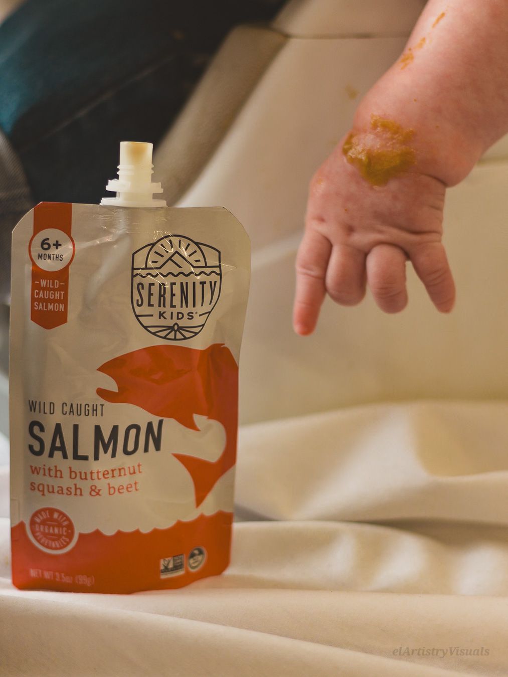Serenity Kids wild caught salmon baby food pouch next to a baby hand covered in orange puree.