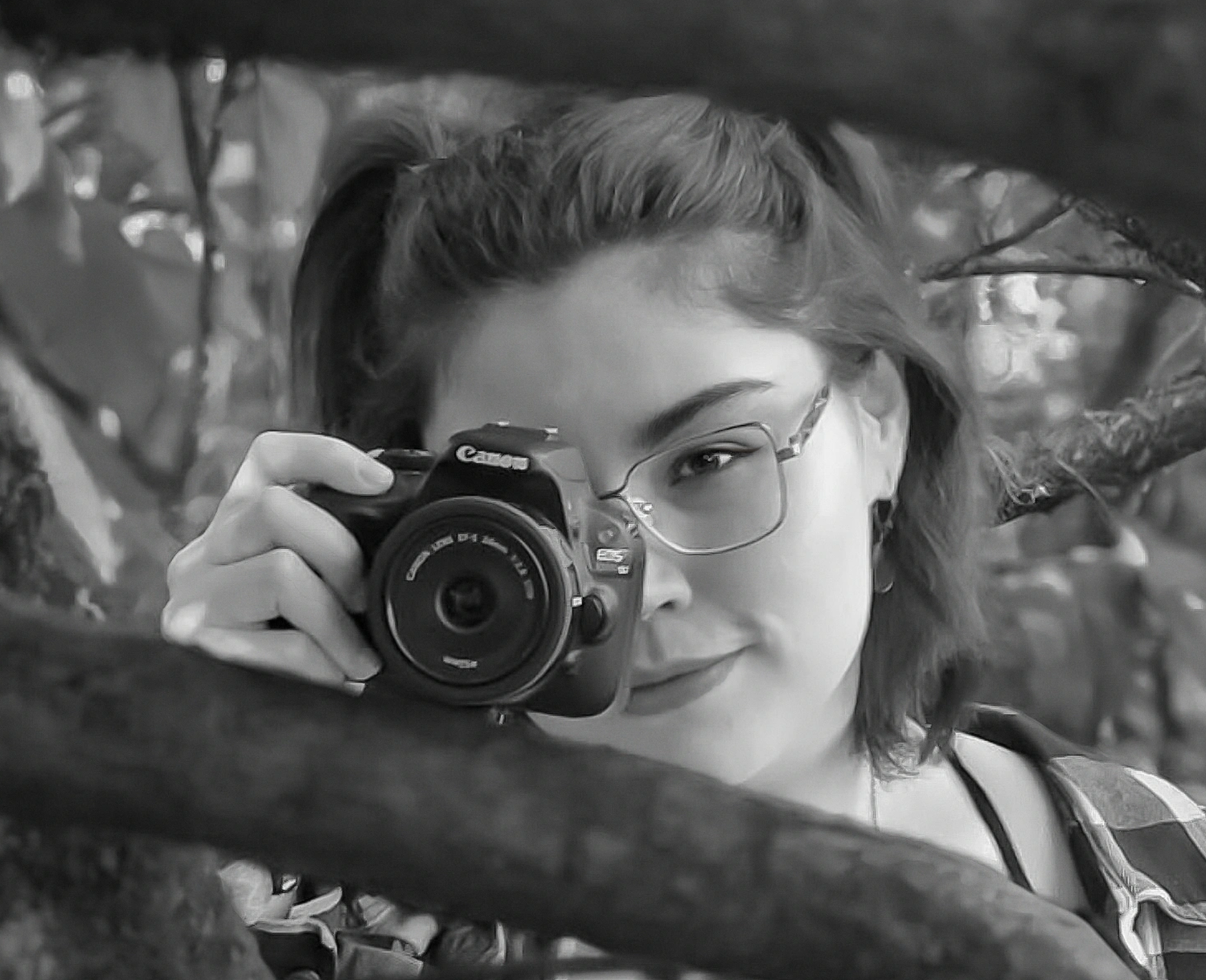 Close-up black and white portrait of a smiling woman with glasses holding a Canon camera among tree branches.