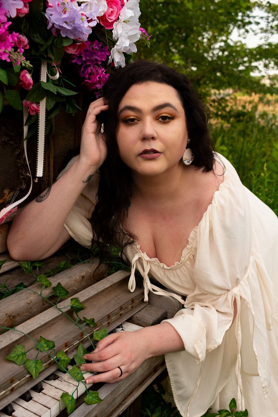 A brunette woman in a cream off-shoulder dress poses by a rustic piano surrounded by colorful flowers outdoors.