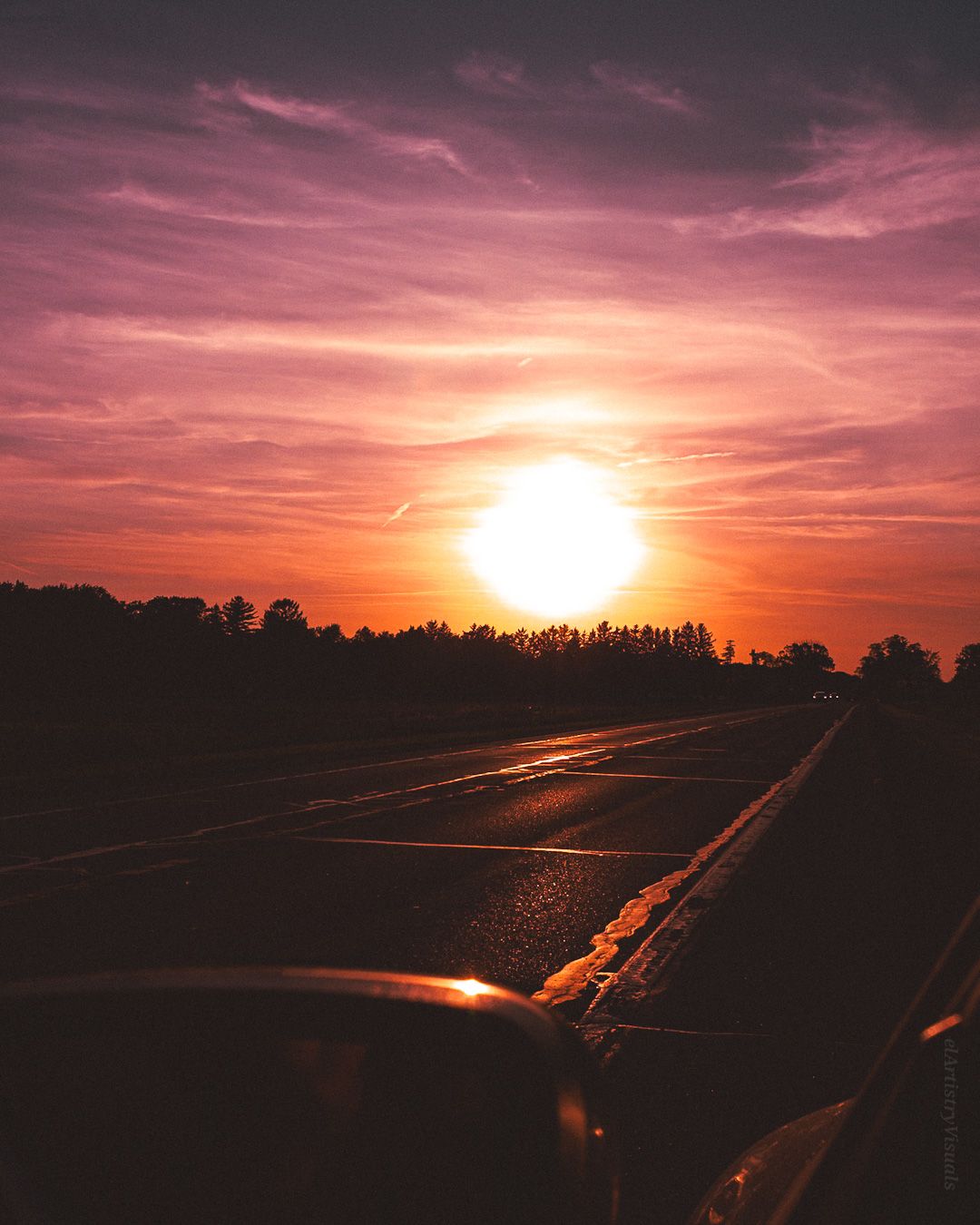 Dramatic orange and purple sunset over a wet highway with silhouetted trees on the horizon.