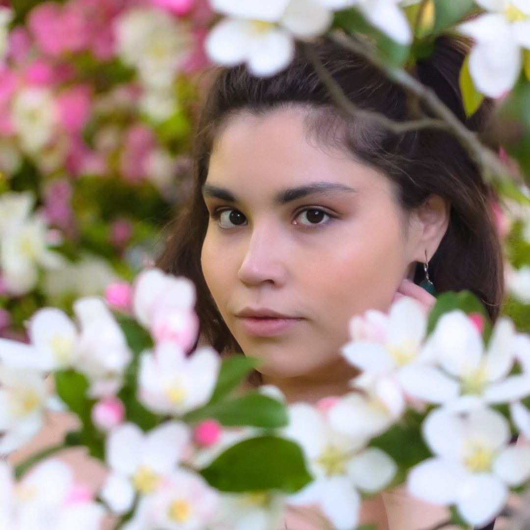 A young woman peers through blooming pink and white apple blossoms in a serene spring garden portrait.
