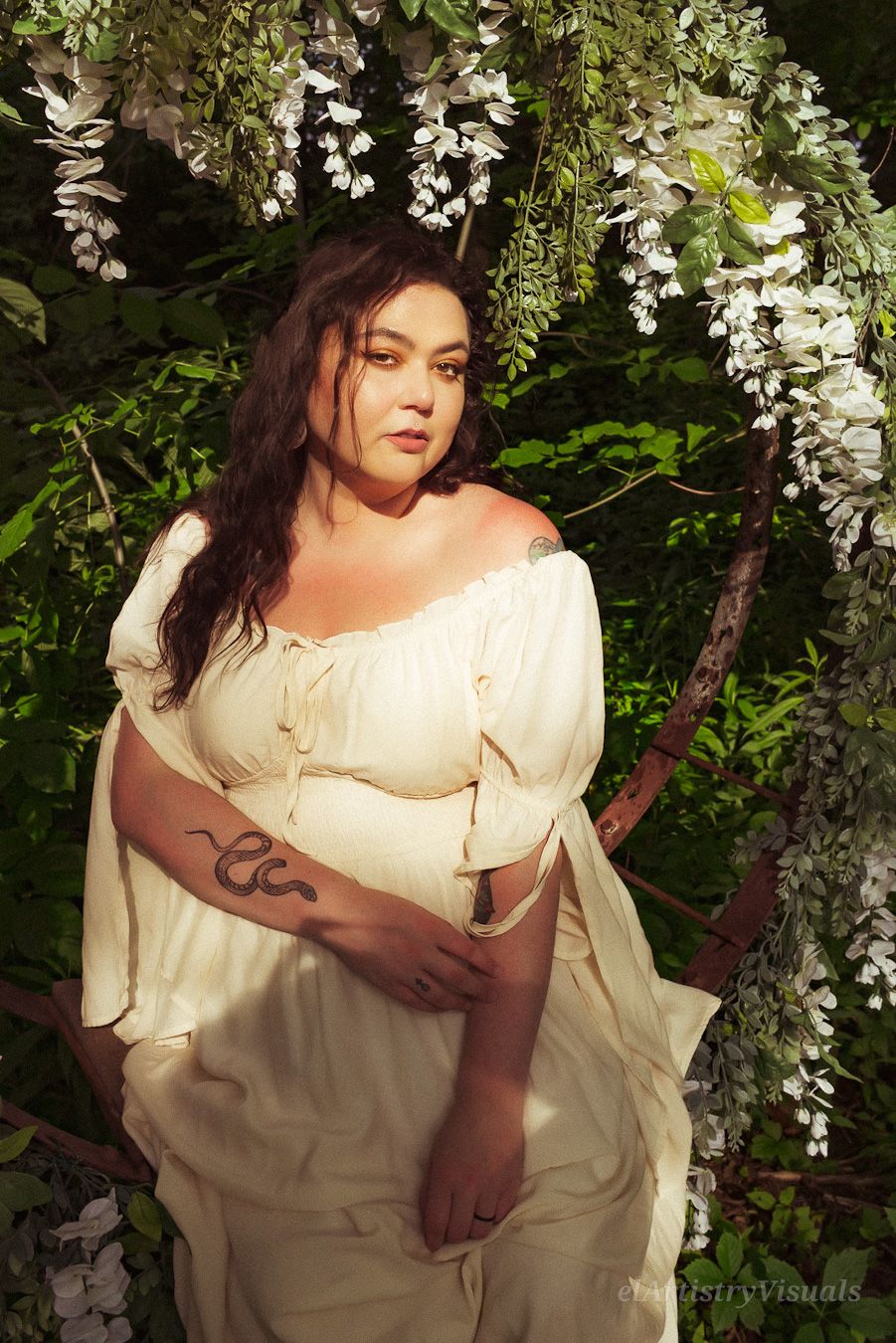 A plus-size woman in a cream off-shoulder dress poses under white wisteria flowers in sunlight.