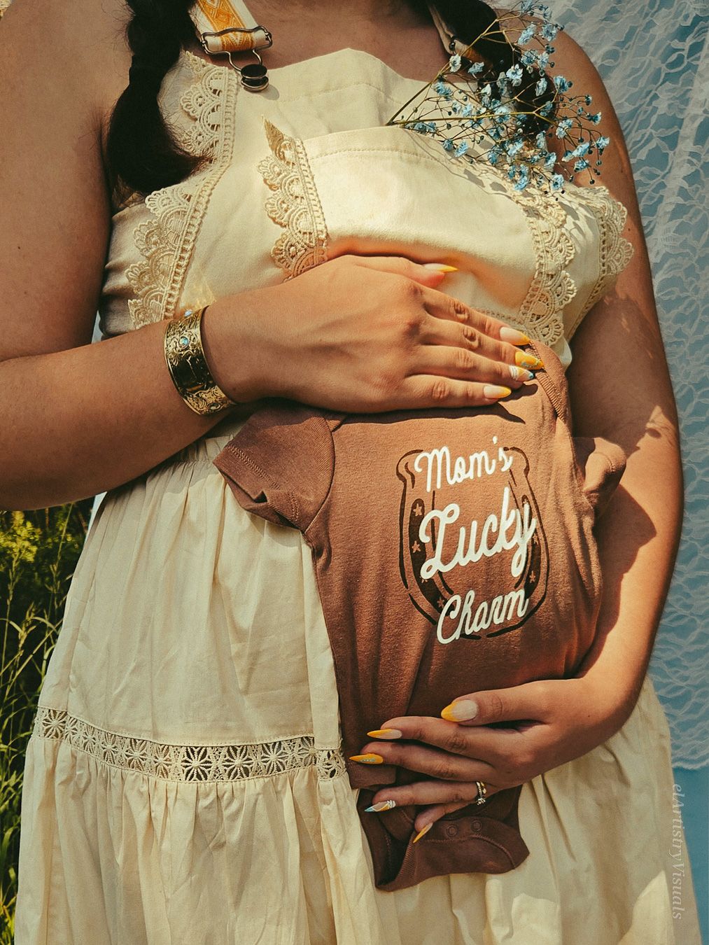 A pregnant woman in a lace dress cradles her baby bump wearing a Mom's Lucky Charm onesie.