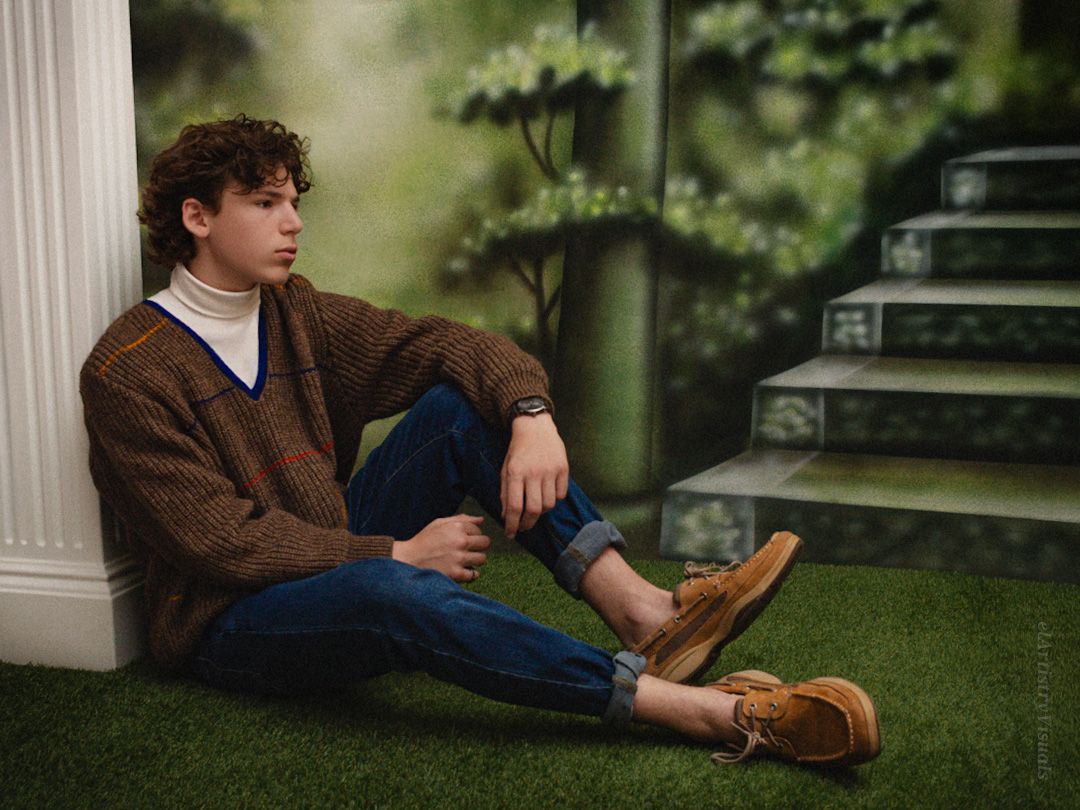 Young man in brown cardigan and jeans sitting against wall with painted forest backdrop.