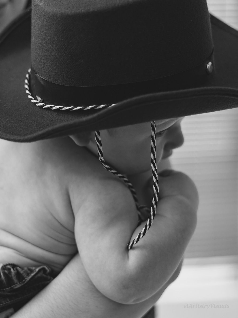 Close-up black and white photo of a baby wearing an oversized cowboy hat, grasping the hat cord.