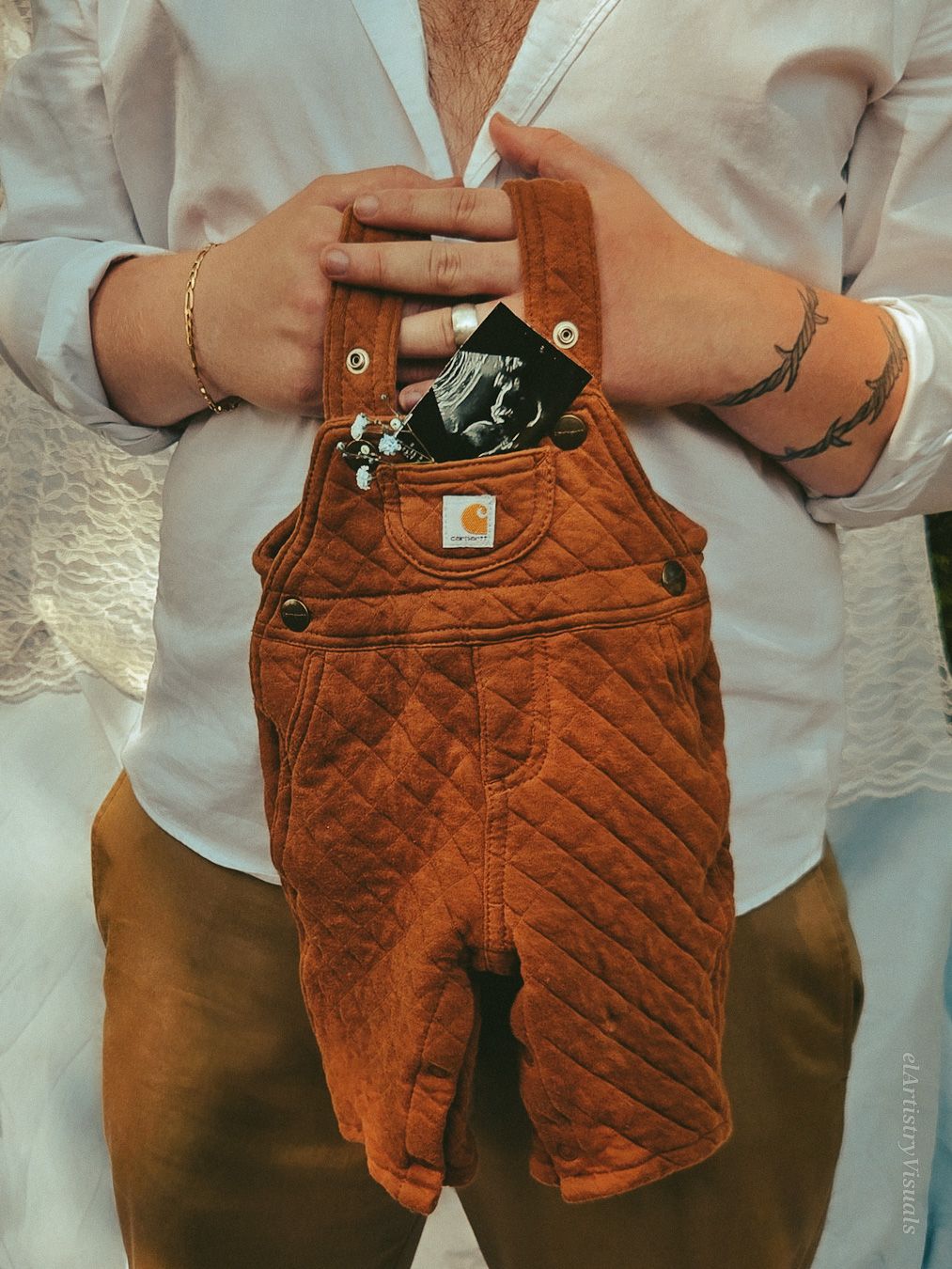 Close-up of tattooed hands holding brown Carhartt baby overalls with a ultrasound photo tucked inside the pocket.