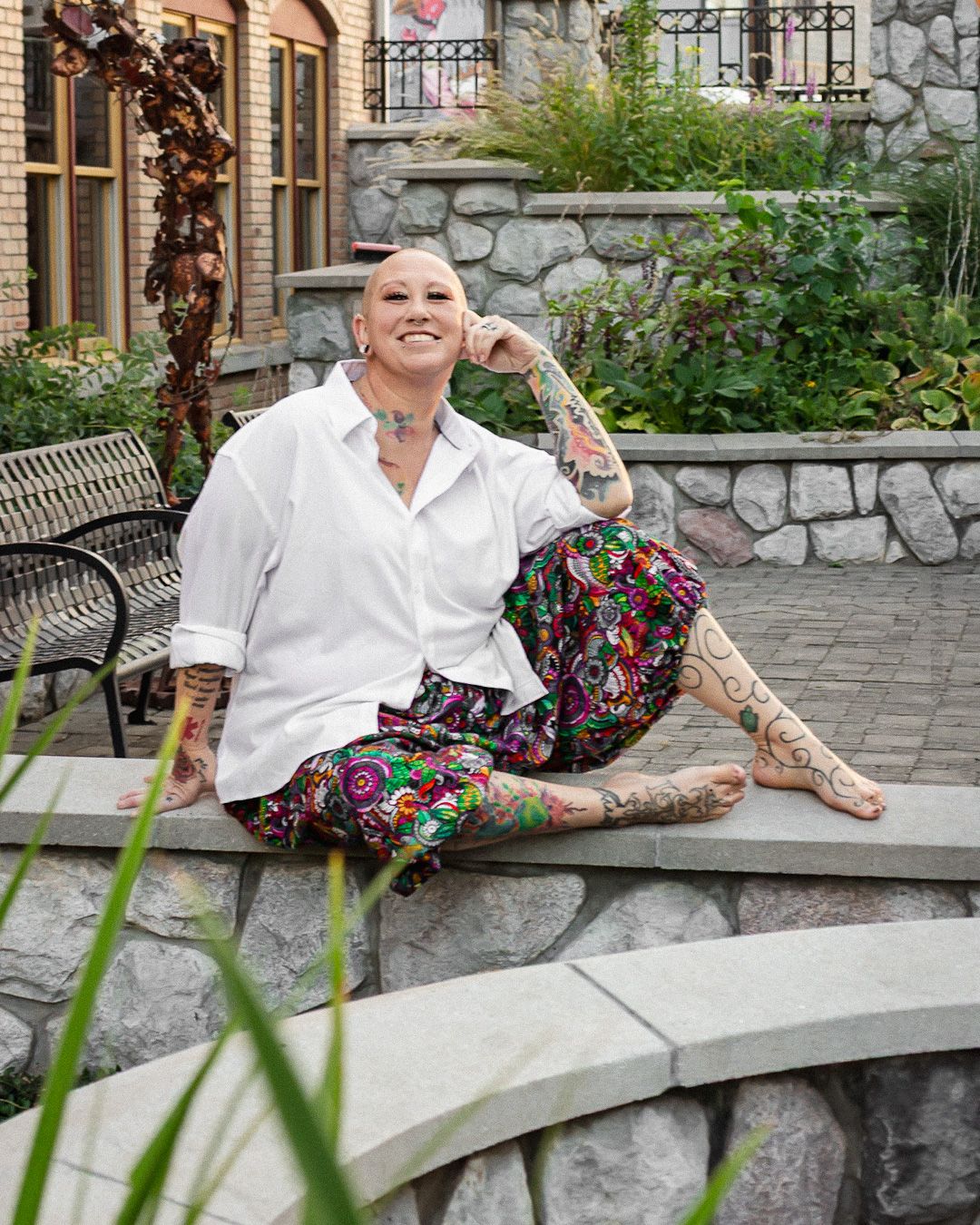 A bald tattooed woman smiles while sitting on a stone wall in colorful pants at an outdoor garden setting.