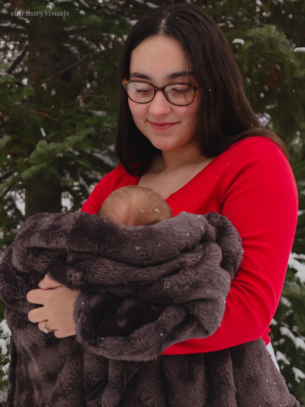 A young mother in a red sweater smiles while holding her newborn wrapped in a gray blanket in snow.