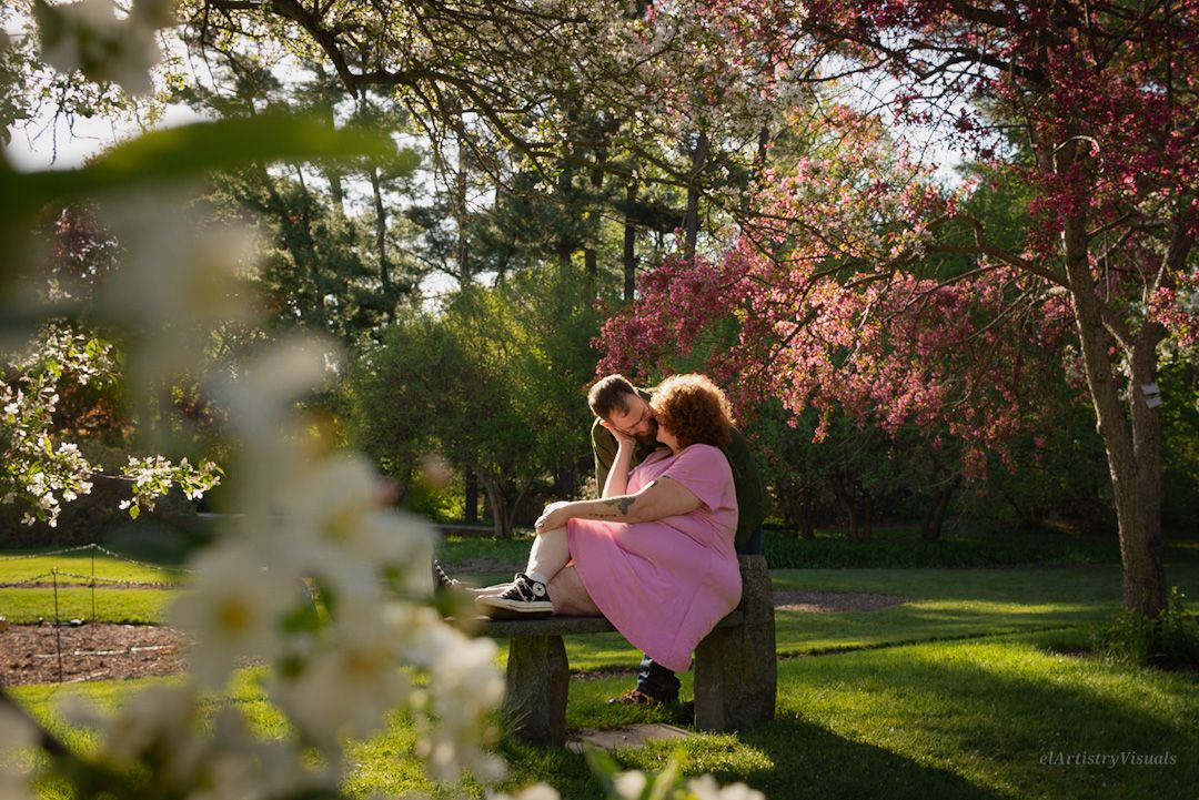 Two people sit closely on a bench in a blooming spring park with pink and white flowering trees.