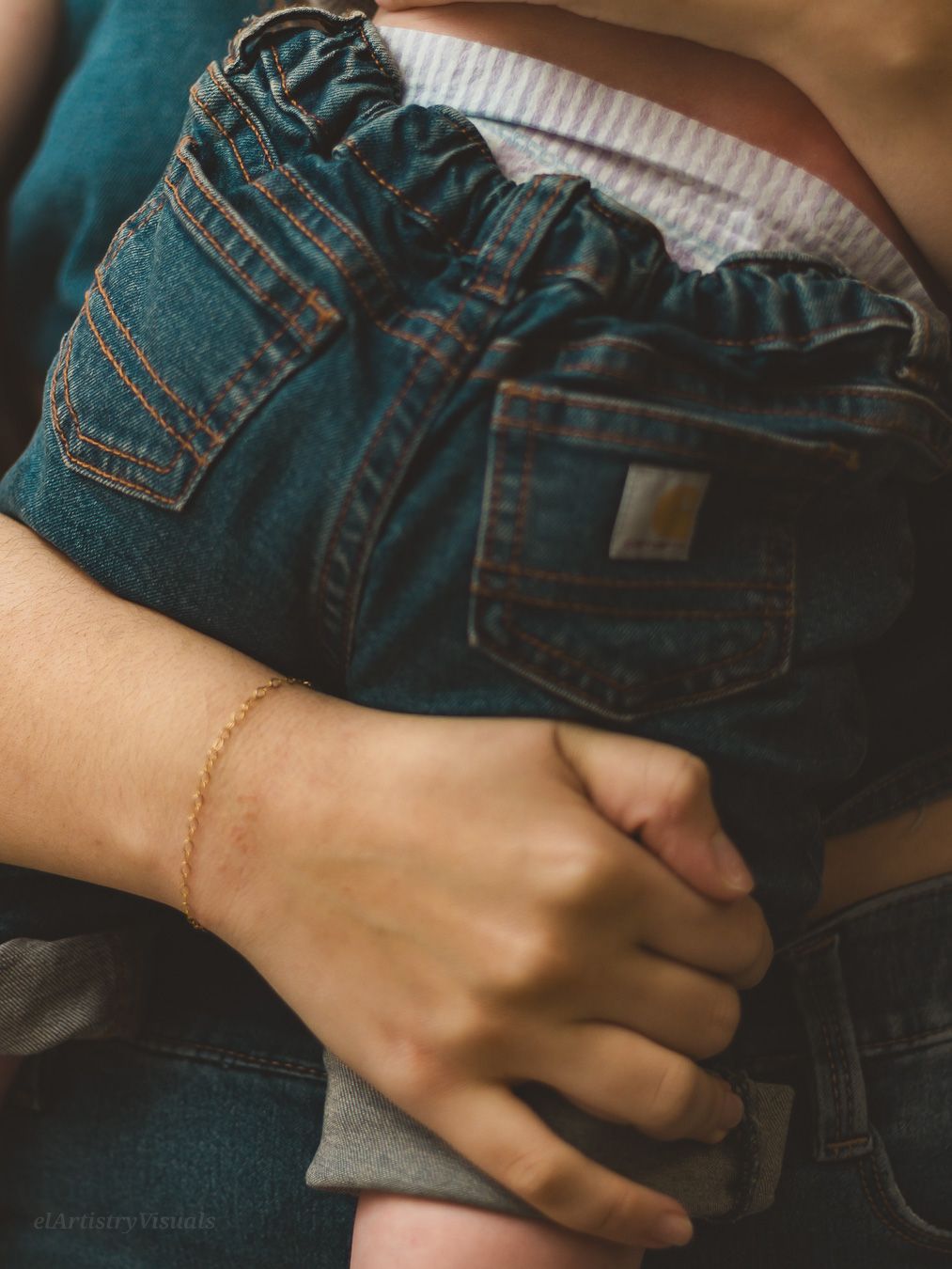 Close-up of a mother's hand with a delicate gold bracelet wrapped around a baby in denim pants.