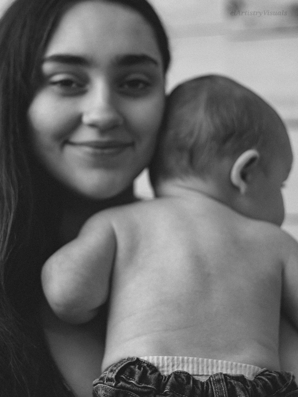 Black and white portrait of a young woman smiling while holding a newborn baby over her shoulder.