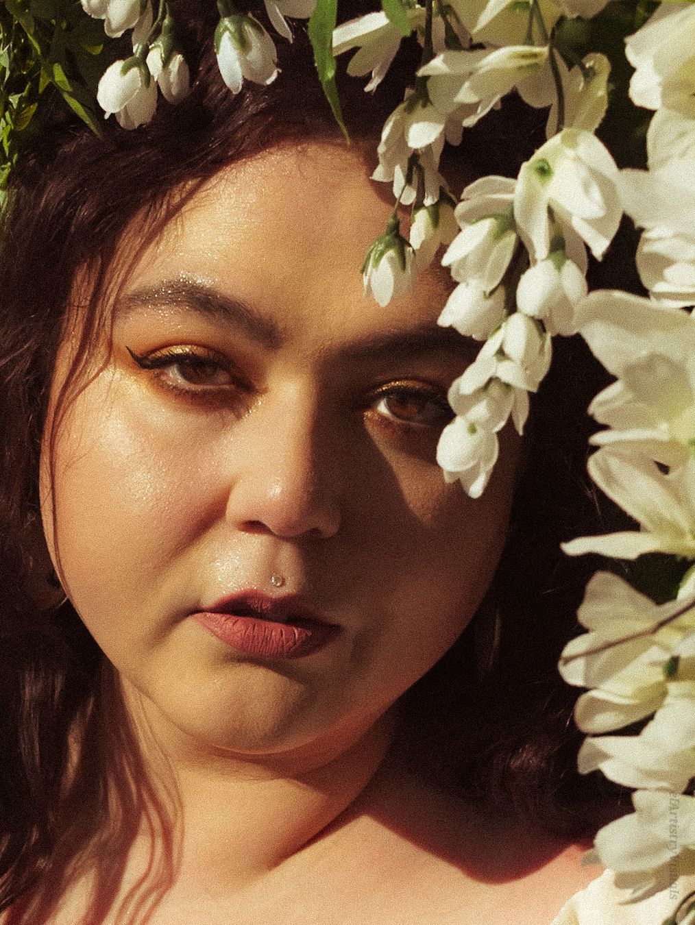 Close-up portrait of a woman with a nose ring surrounded by white wisteria flowers in warm sunlight.