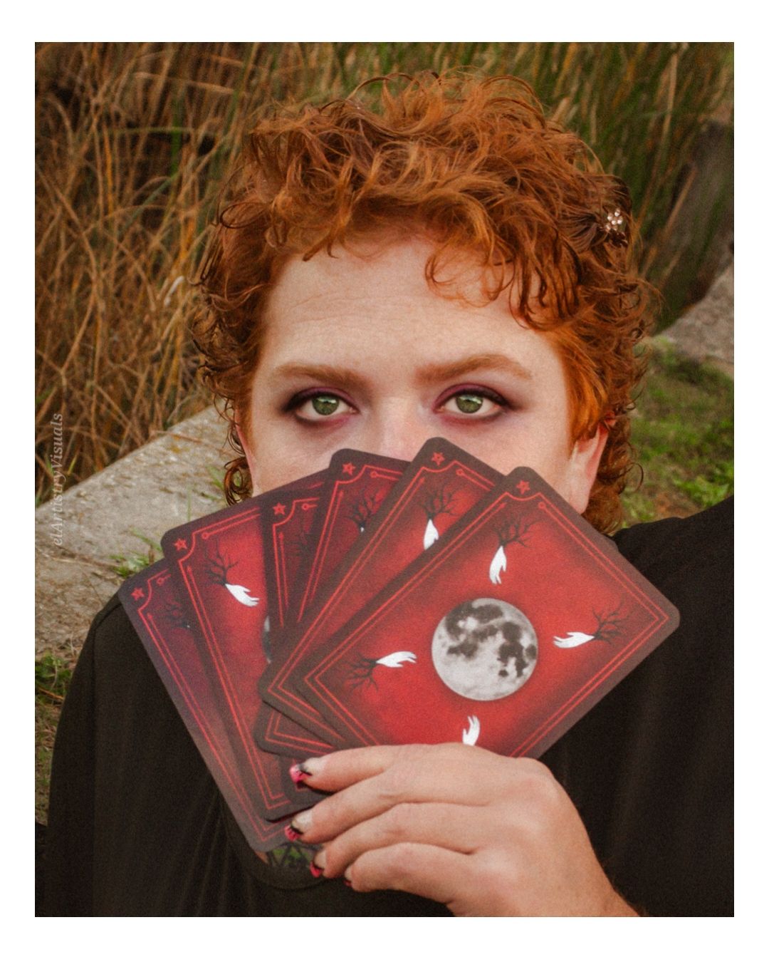Woman with curly red hair holding a fan of red moon-themed playing cards over her face outdoors.