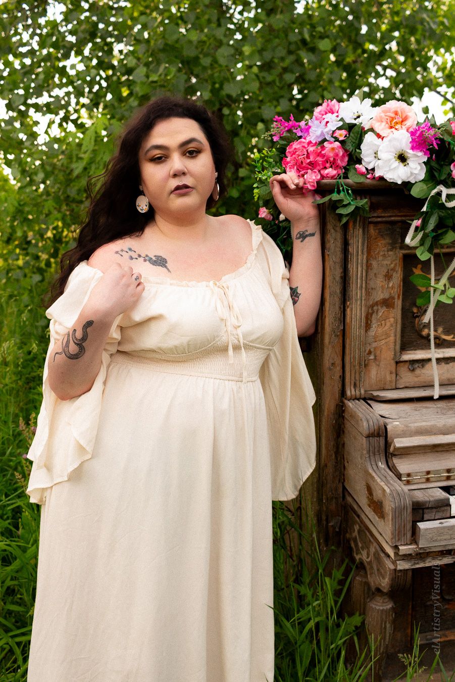 A tattooed woman in a cream Renaissance-style dress stands beside a rustic wooden dresser with flowers.