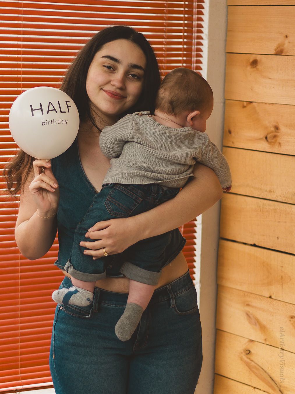 A young mother holds her baby and a half birthday balloon while standing near wooden wall indoors.