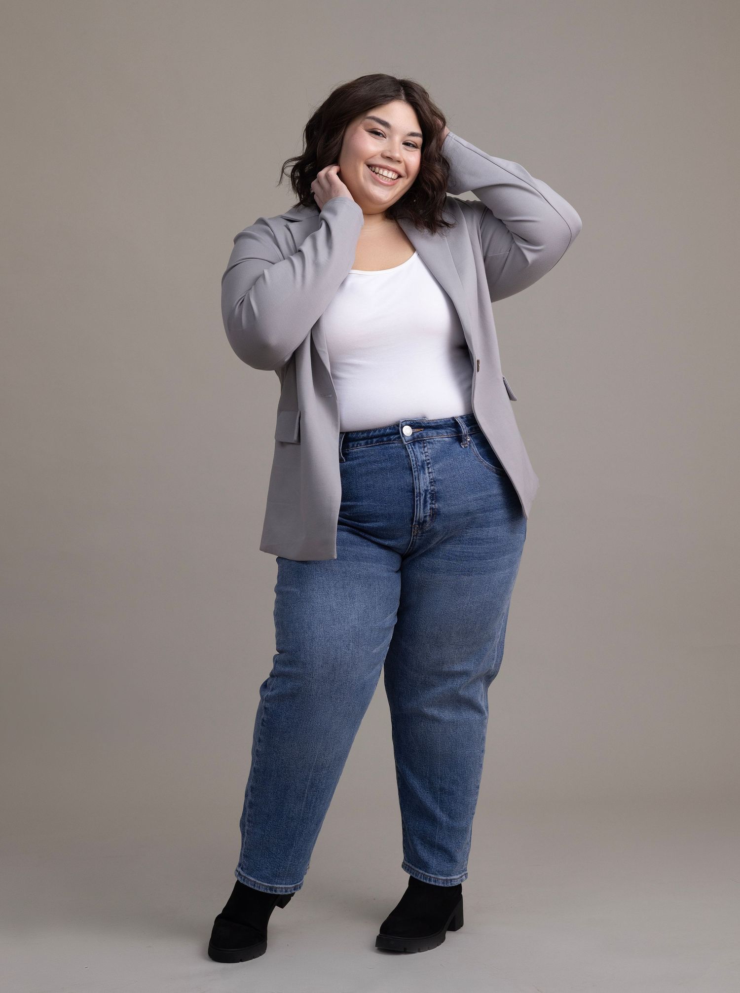 A plus-size woman smiles confidently in a gray blazer, white top, and blue jeans against a neutral background.