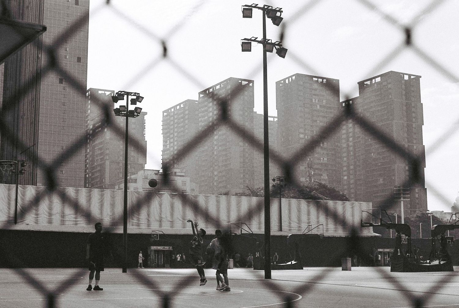 Black and white photo of people playing basketball on an urban court, viewed through a chain-link fence with city skyscrapers behind.