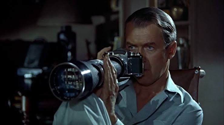 Man in white shirt peers through a large telephoto camera lens in a dimly lit room.