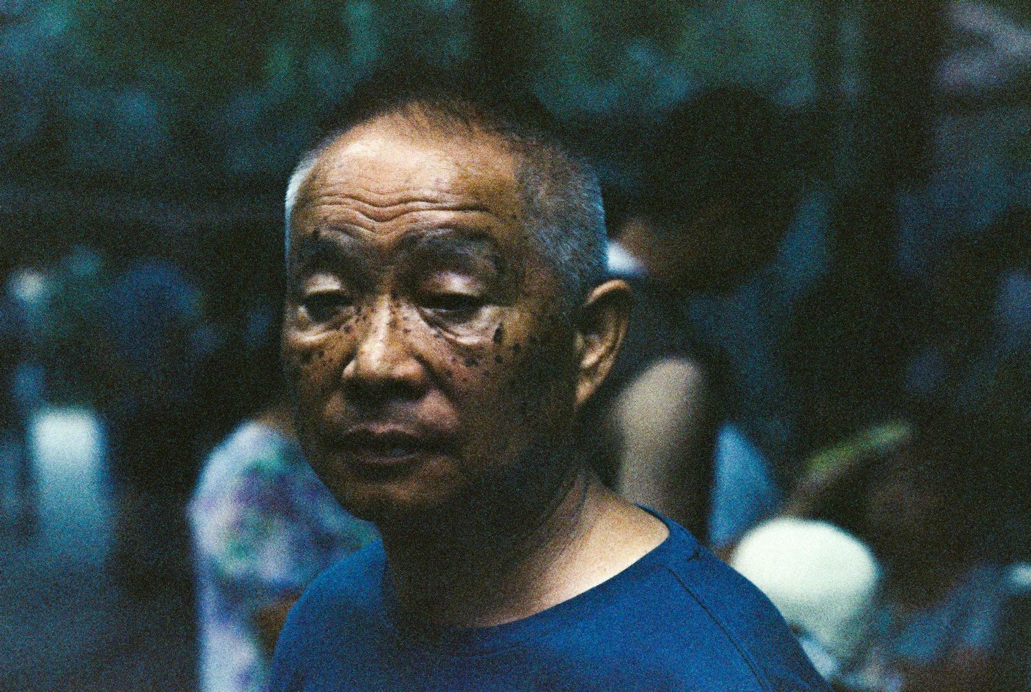Close portrait of an elderly Asian man in a blue shirt with a crowd blurred in the background.