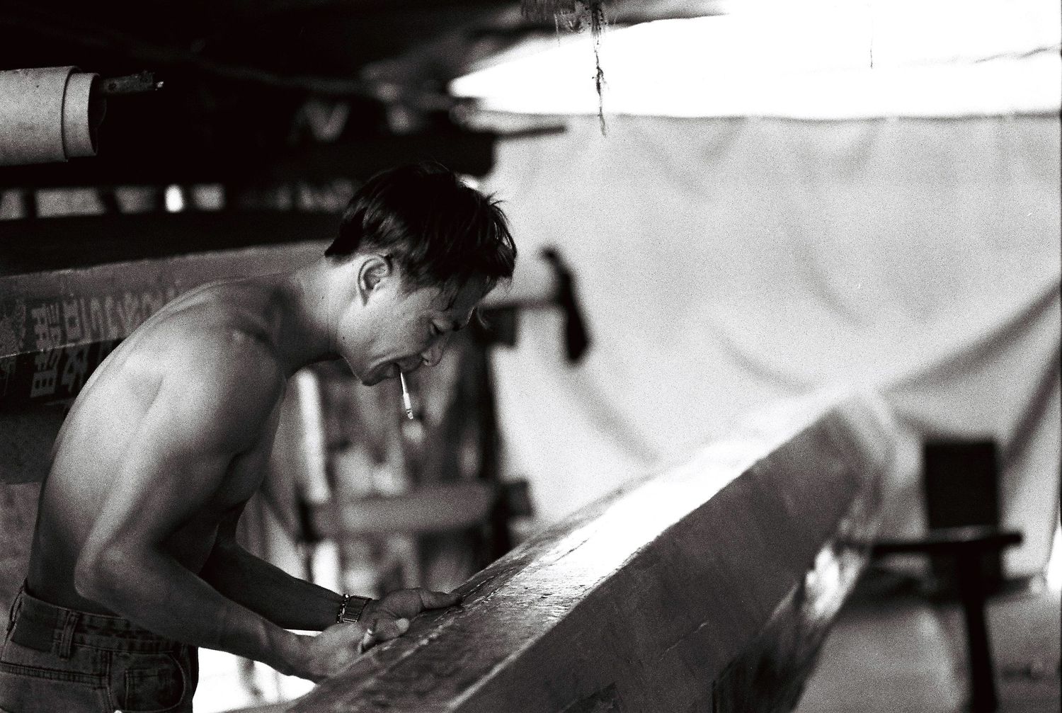 Black and white photo of a shirtless man shaping a surfboard in a workshop in Foshan, China.