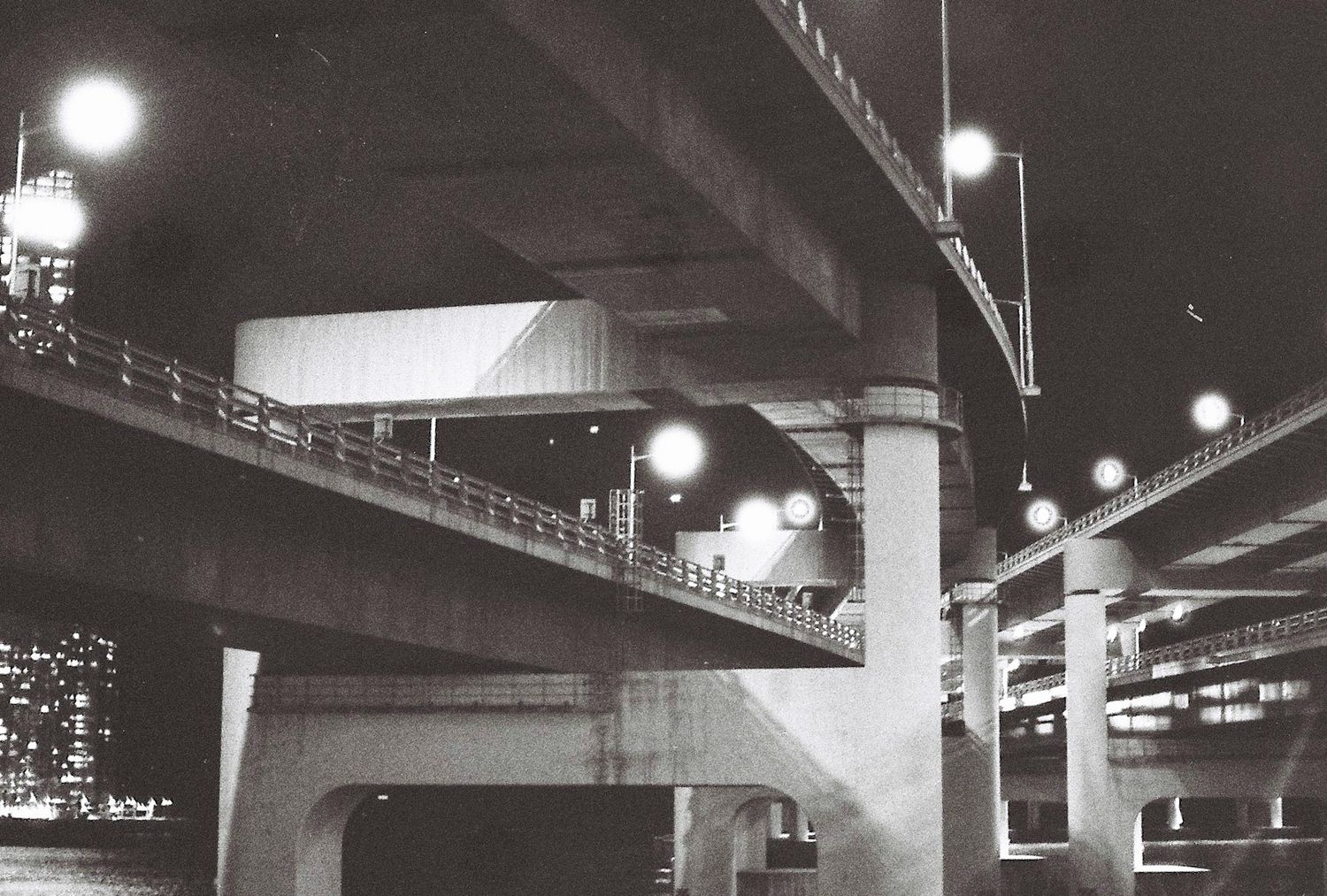Black and white night photo of illuminated urban highway overpass with glowing street lamps and city lights in background.