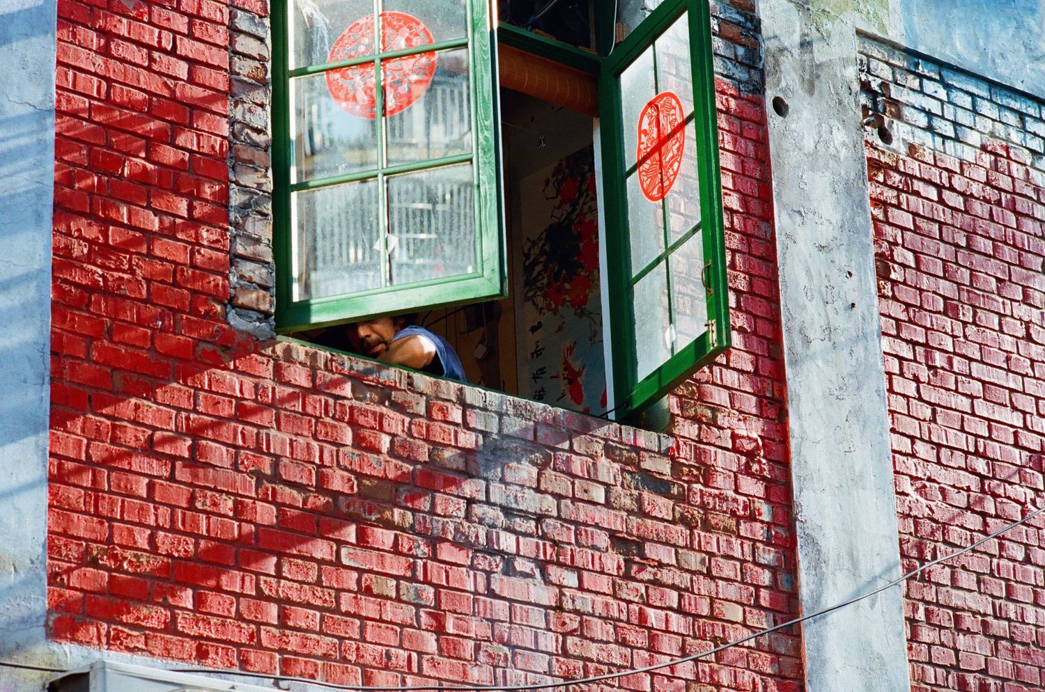 Person leaning out of green-framed window on red brick building with decorative murals.