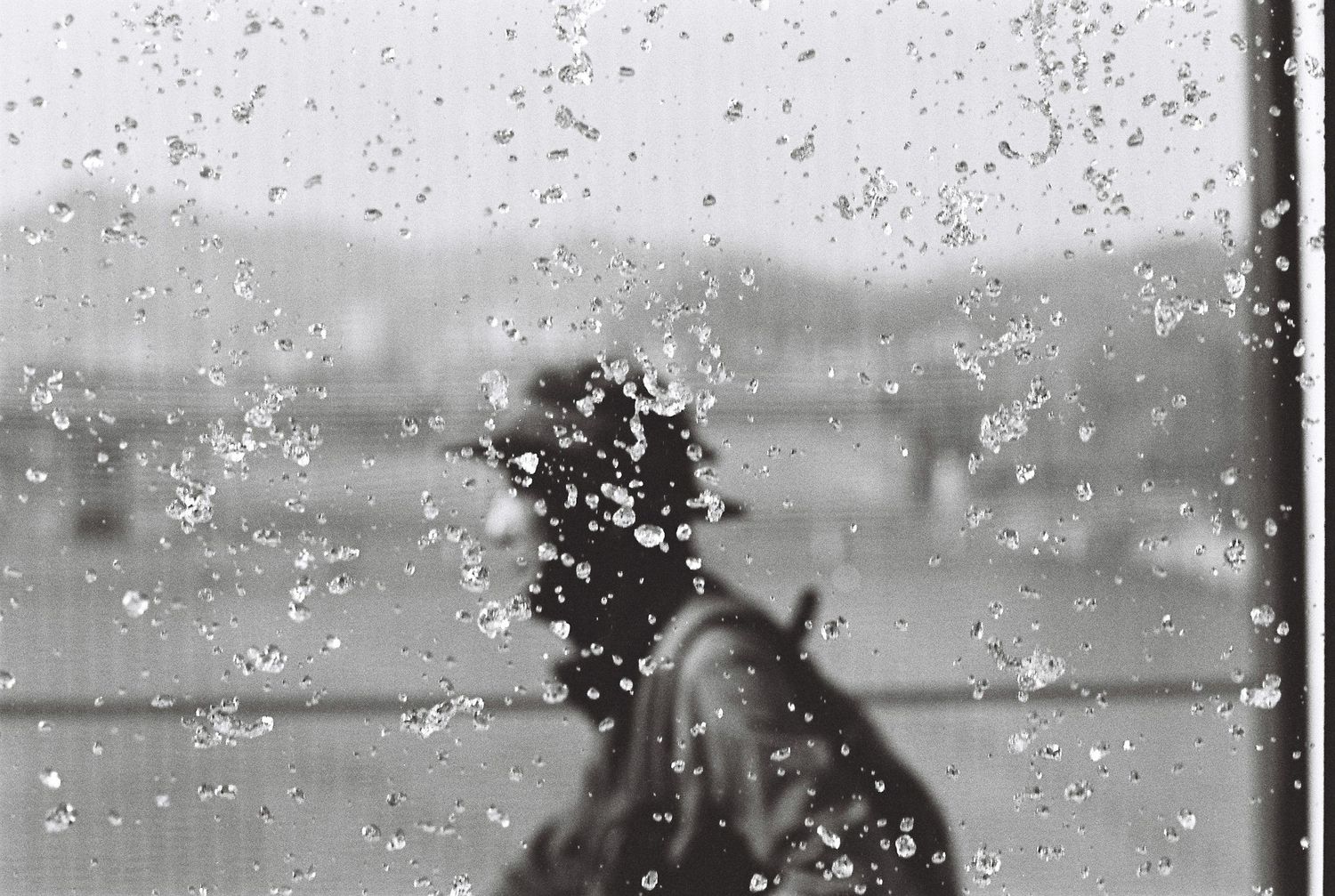 Raindrops on a window glass with a blurred figure visible through the wet pane in black and white.