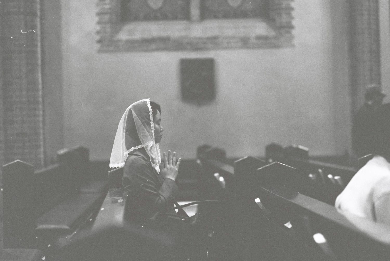A woman in a white headscarf kneels in prayer alone in a dimly lit church pew in Seoul.