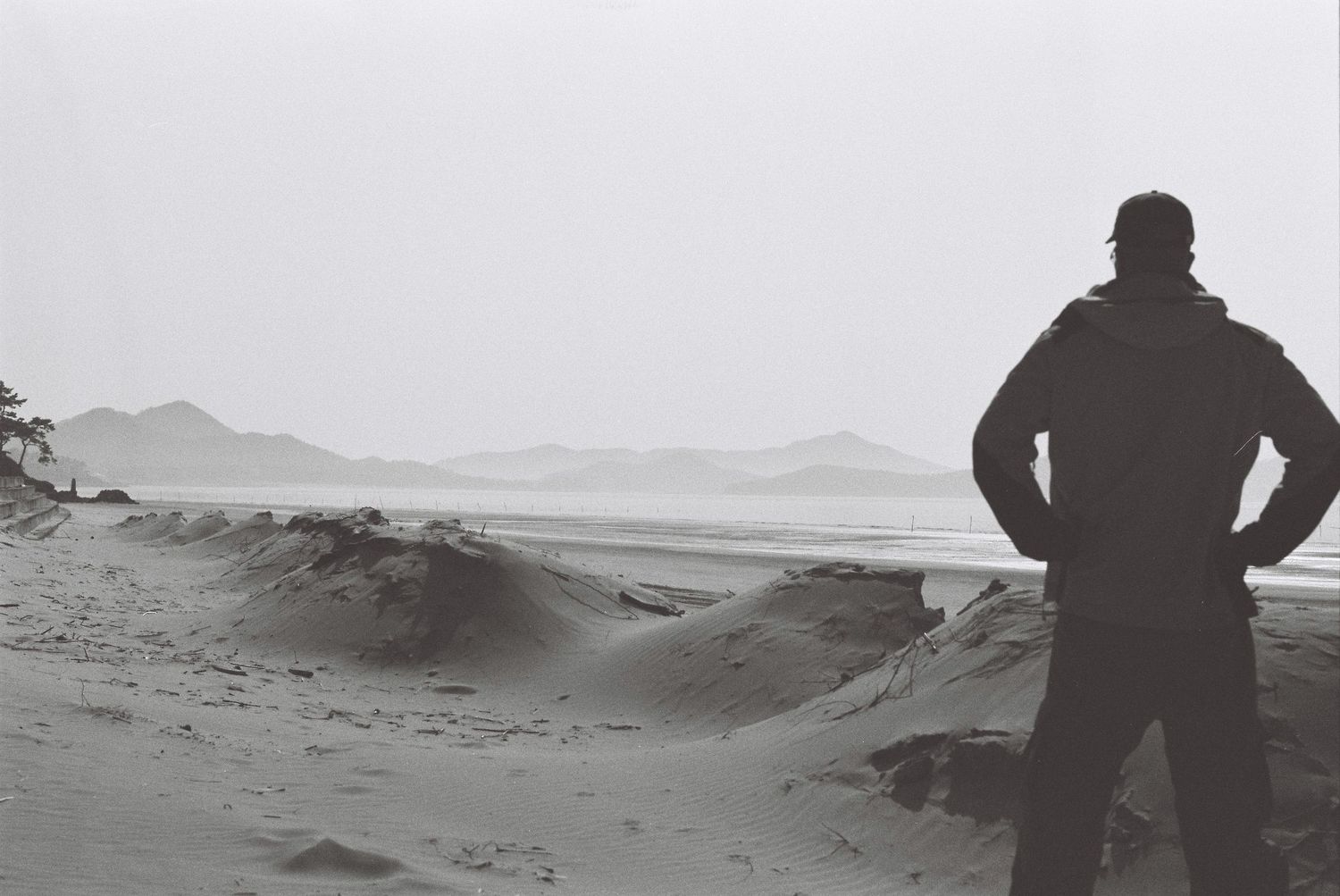 Solitary figure stands on a vast sandy beach gazing at misty mountains in a black and white landscape photo.