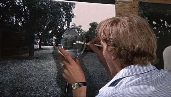 Person examining a black and white photograph through a magnifying glass outdoors near trees.