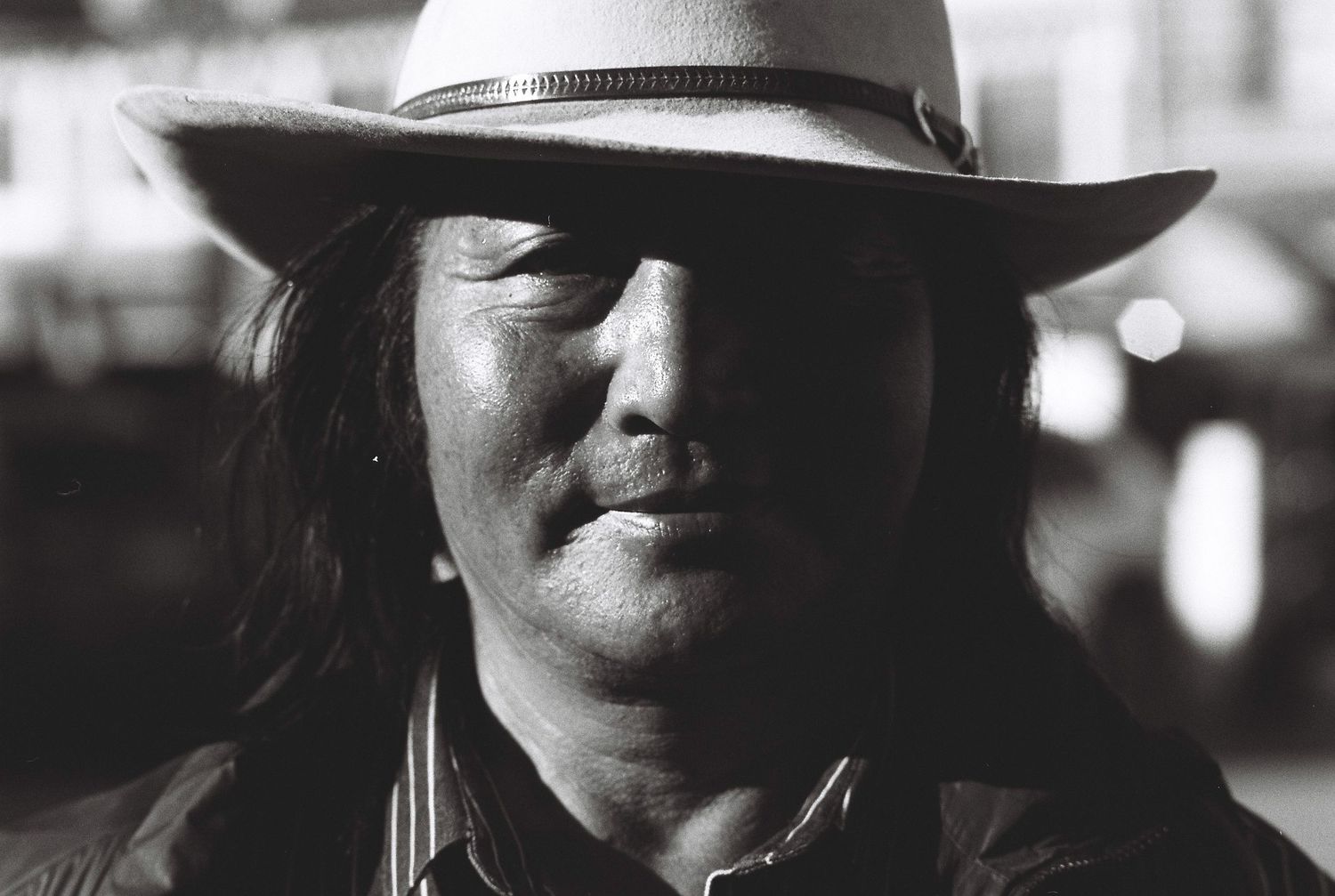 Black and white portrait of a chinese man with long hair wearing a white cowboy hat outdoors.
